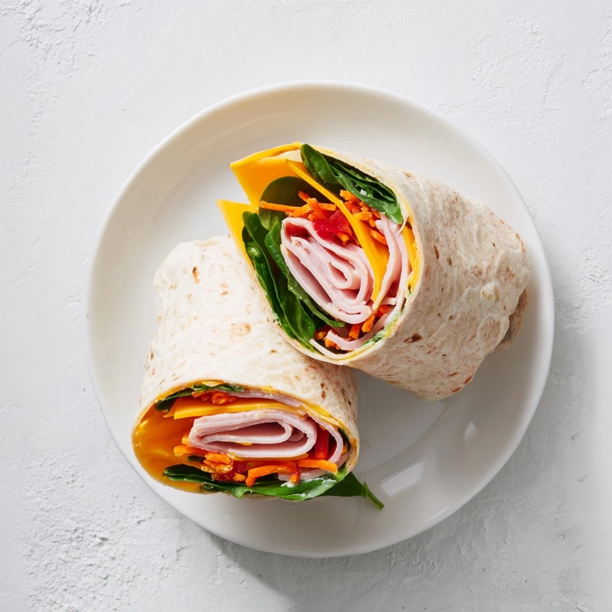 Two halved Turkey Snack Wrap Rolls on a white plate, showcasing layers of turkey, cheddar, spinach, carrots, and red bell pepper.