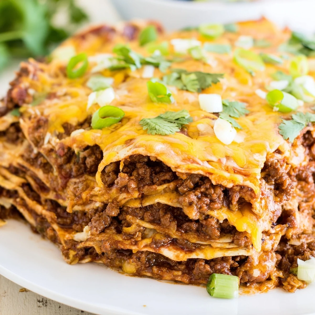 A close-up view shows a hearty slice of Beef Enchilada Casserole on a white plate, garnished with fresh cilantro and green onions for a pop of color. 