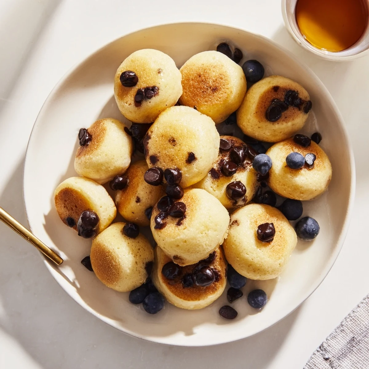 Bite-sized Pancake Poppers baked in a mini muffin tin, filled with blueberries and served alongside a small cup of yogurt.