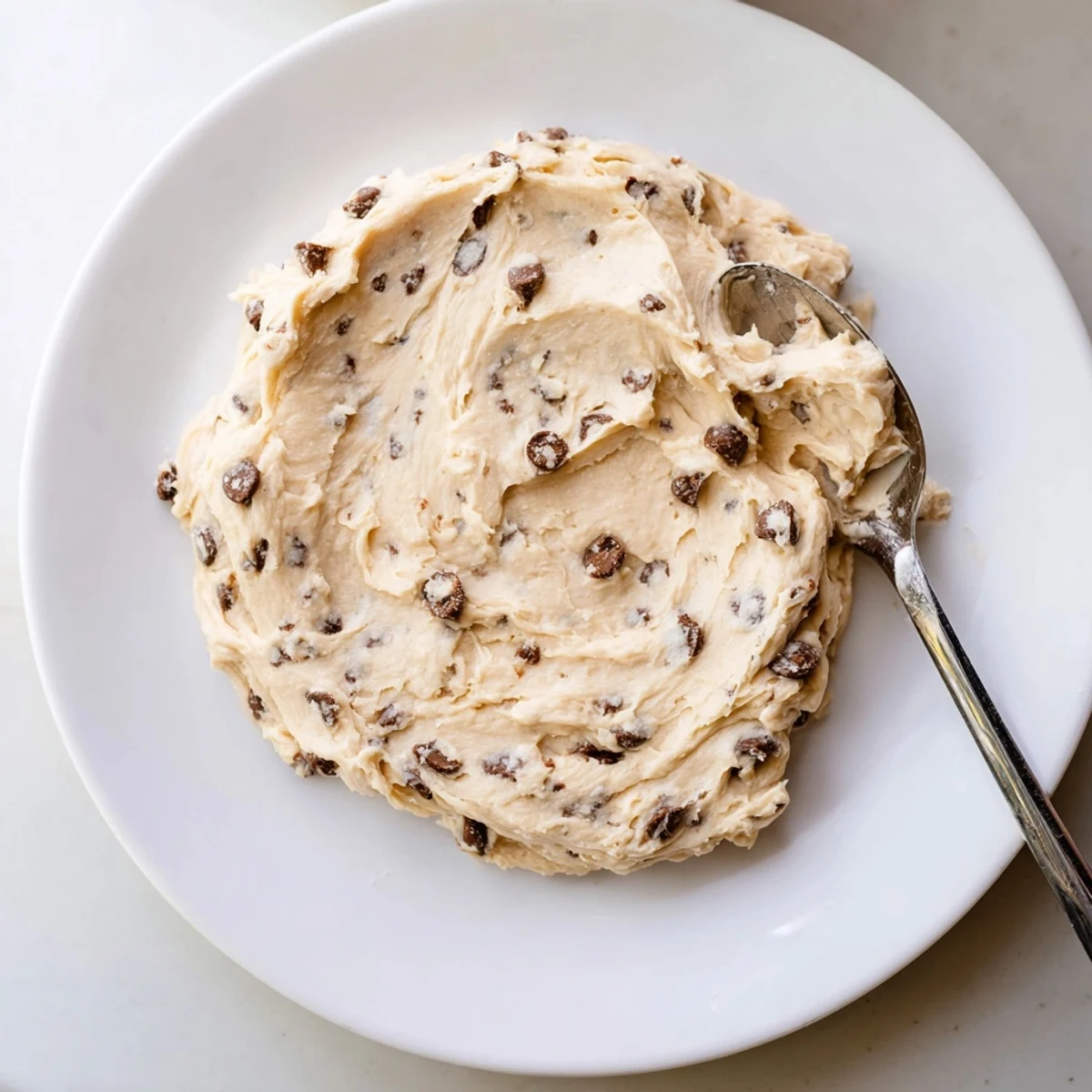 Creamy Cookie Dough Frosting in a bowl with mini chocolate chips and a wooden spoon, ready to spread on cupcakes.