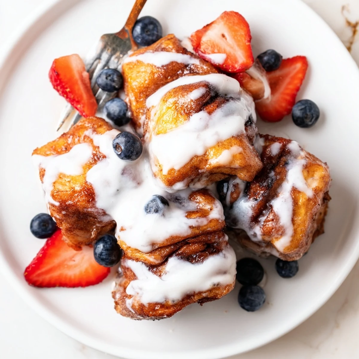 Golden-brown Cinnamon Roll French Toast Bites are drizzled with warm icing on a plate, served with fresh berries and maple syrup for a sweet breakfast.