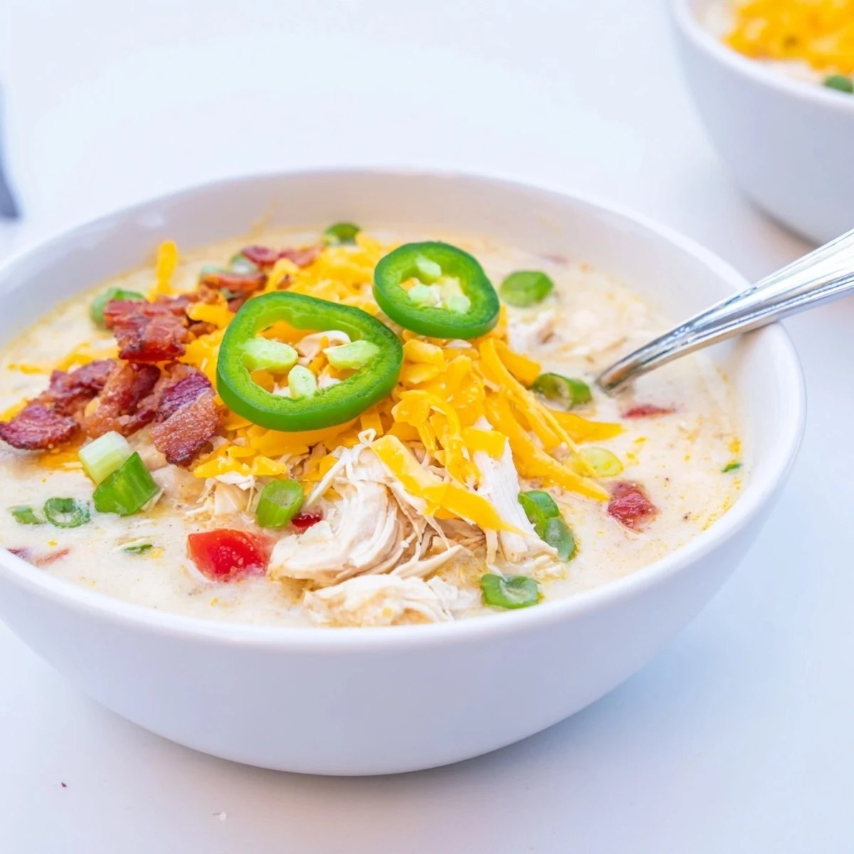 Creamy spicy jalapeno popper chicken soup topped with crumbled bacon and melted cheddar cheese in a white bowl